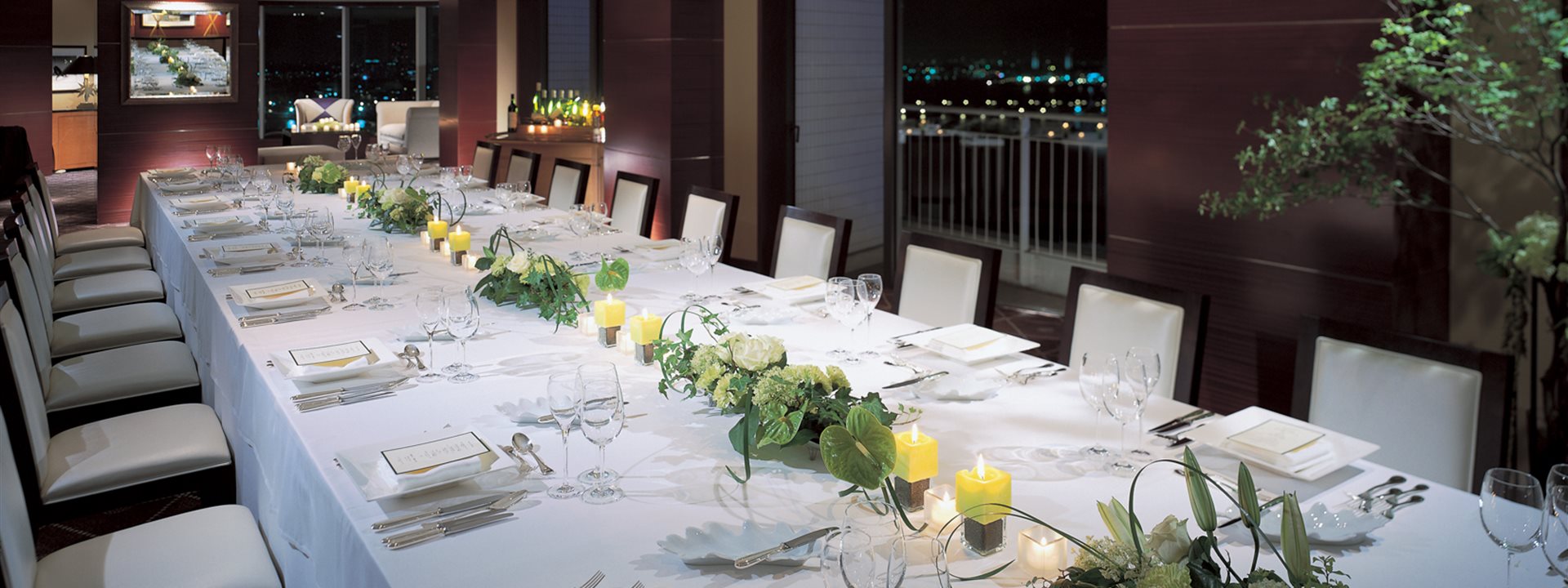 A long table is set up for a dinner party in the "Pacific Room" on the top floor of the Yokohama Bay Hotel Tokyu. The night view stretches out in the background.